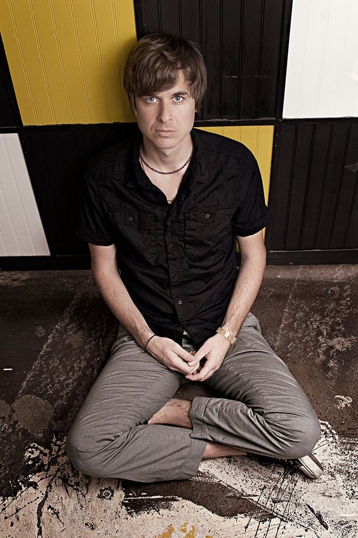David Dewese sits cross-legged on a paint-spattered floor in a black shirt, in front of black, white and yellow wall panels.