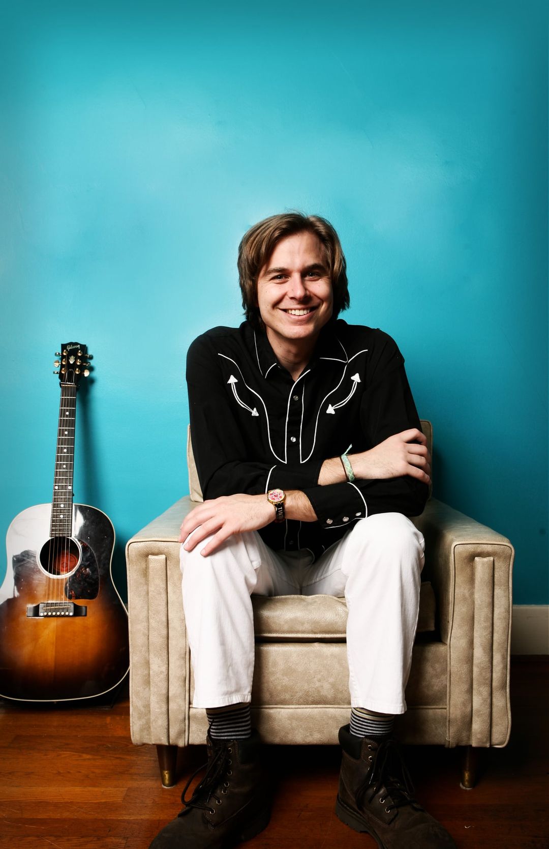 David Dewese smiles from a cream armchair against a turquoise wall, in a black western shirt and white jeans, with an acoustic guitar leaning beside him.