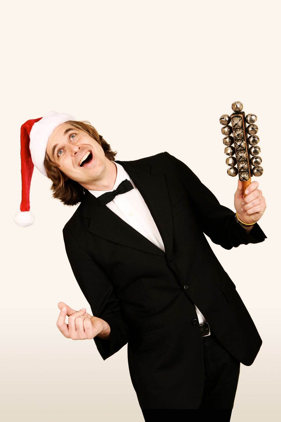 David Dewese in a tuxedo and Santa hat sings, shaking sleigh bells mounted on a wooden handle, against a plain cream backdrop.