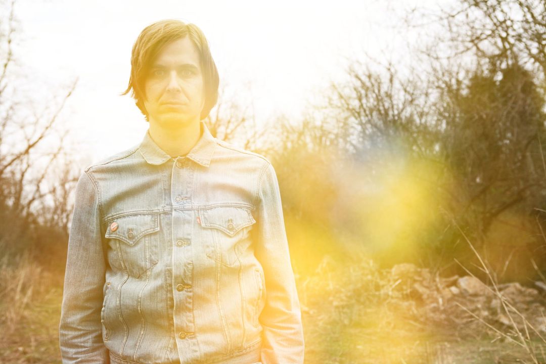 David Dewese in a denim jacket stands in bare woodland, backlit by hazy golden sunlight.