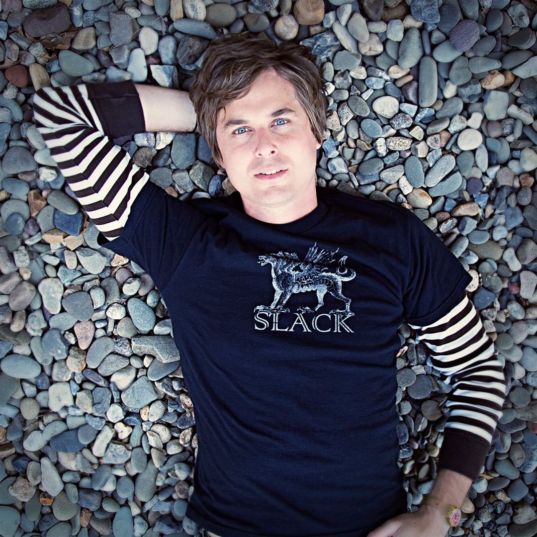 David Dewese lies on smooth river stones, one arm behind his head, in a navy SLACK T-shirt over a striped long-sleeve shirt.