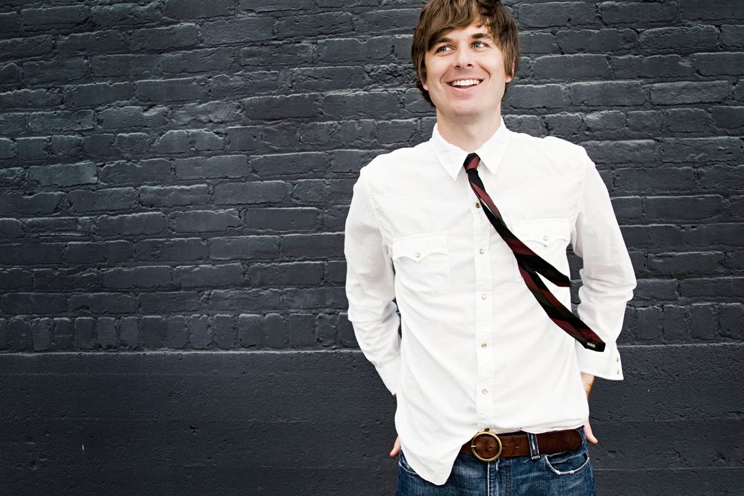 David Dewese smiles and looks upward in a white western shirt and loosened dark-red tie, against a dark brick wall.