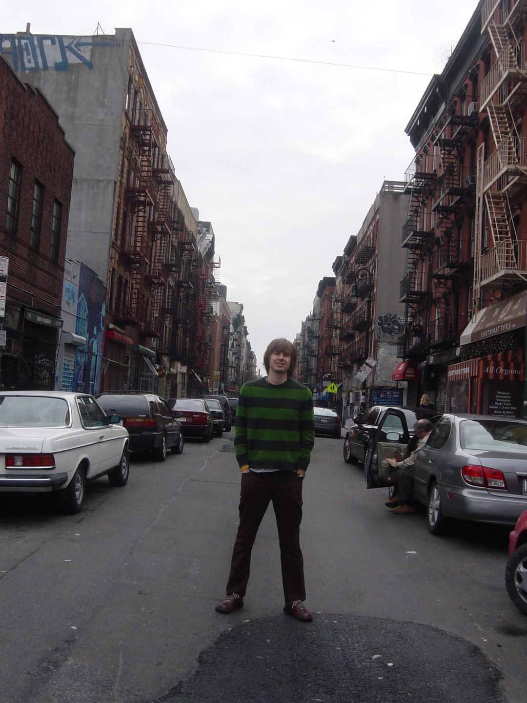 David Dewese, in a green-and-black striped sweater, stands in the middle of a narrow New York City street lined with tenements, fire escapes and parked cars.