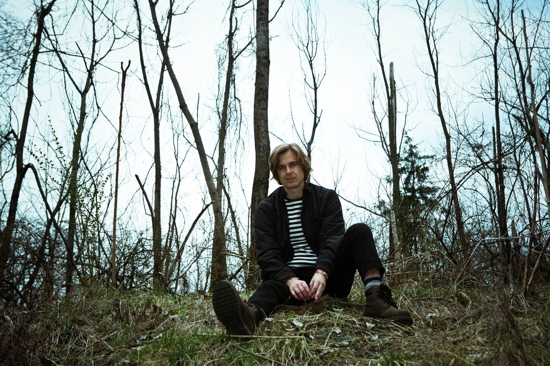 David Dewese, in a black jacket over a striped shirt, sits on a grassy slope among bare saplings under a pale sky.