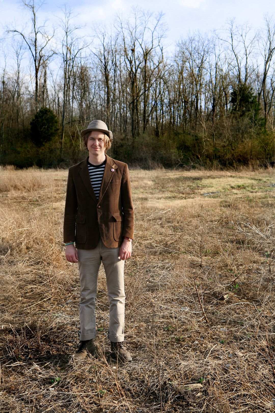 David Dewese, in a felt hat and brown corduroy blazer, stands in a dry winter field in front of bare trees under a blue sky.