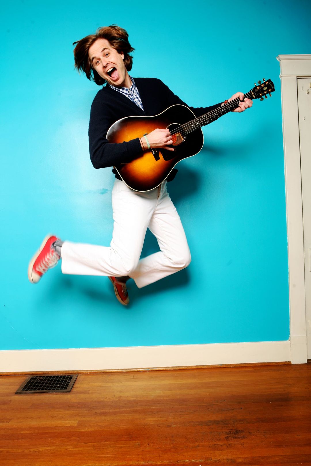 David Dewese leaps in the air with an acoustic guitar against a bright turquoise wall.