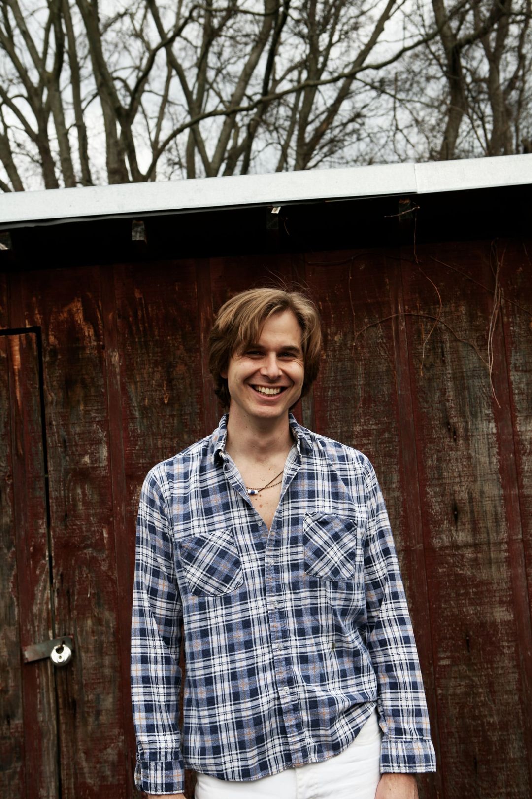 David Dewese smiles in a blue plaid shirt against a weathered red barn wall.