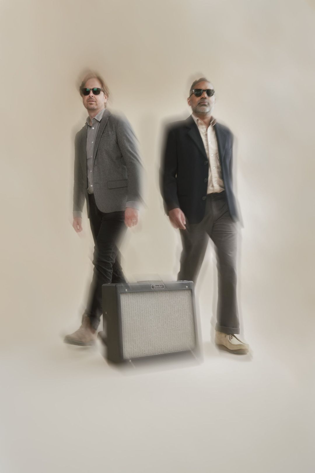 The Foxymorons, David Dewese and Jerry James, walk toward the camera in sunglasses, blurred with motion, a small Fender amp on the floor between them.