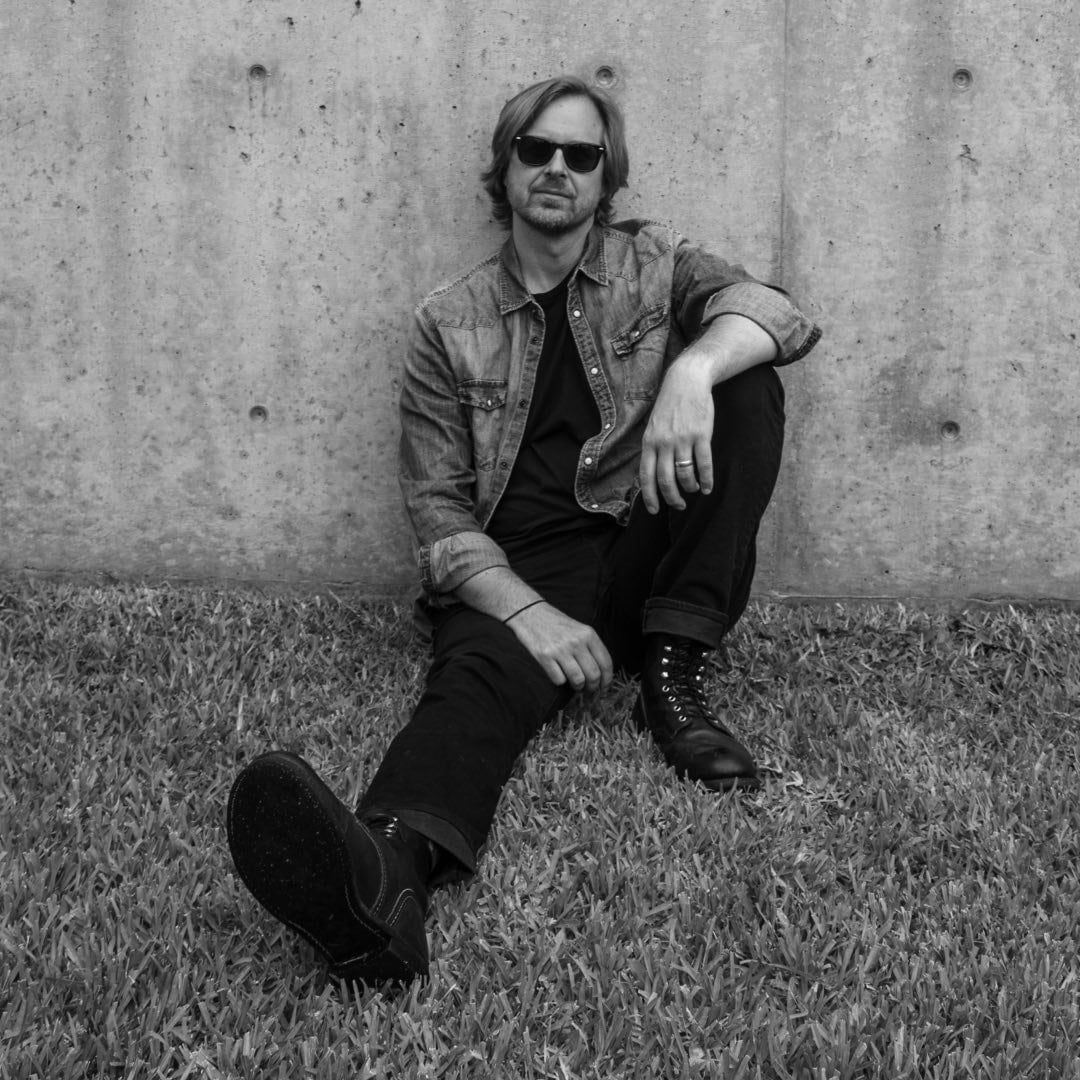 David Dewese in sunglasses and a denim shirt, sitting on grass against a concrete wall, in black and white.