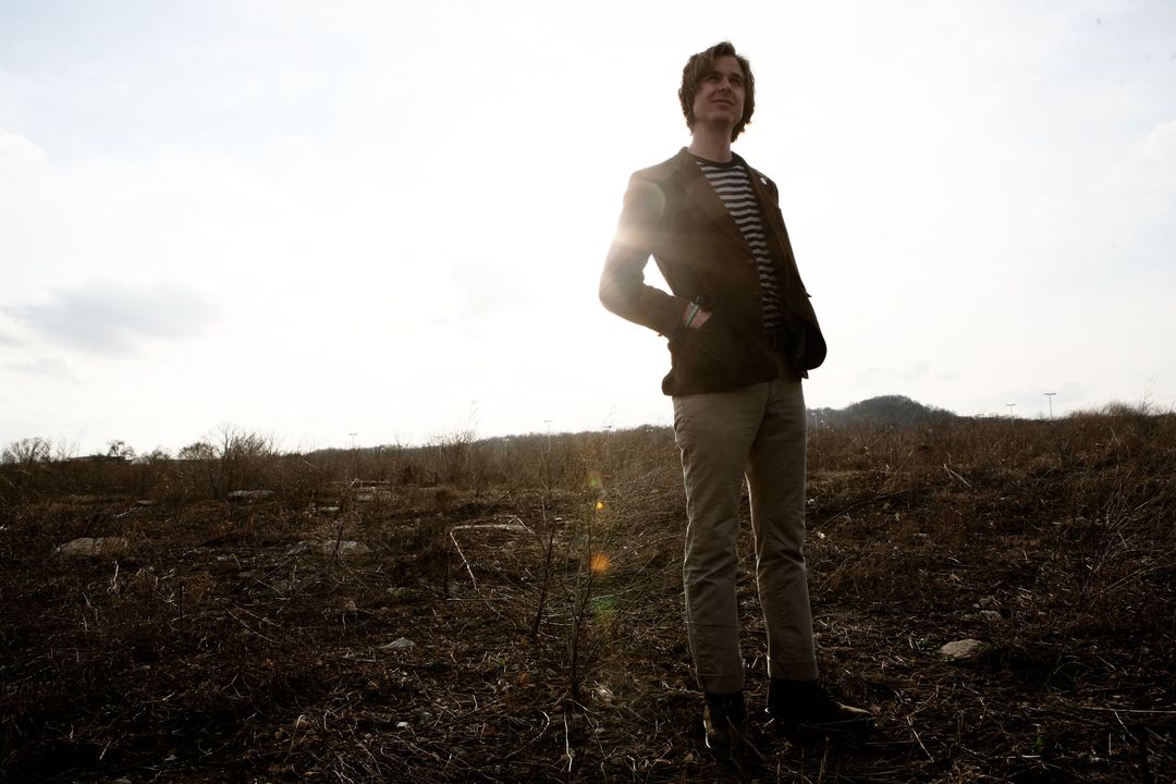 David Dewese stands in a dry, open field under a bright, hazy sky, hands in the pockets of a brown corduroy blazer, gazing up and to the right.
